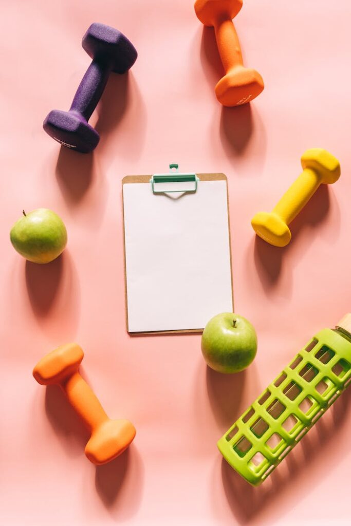 Flat lay of gym weights, apples, clipboard, and water bottle on peach background.